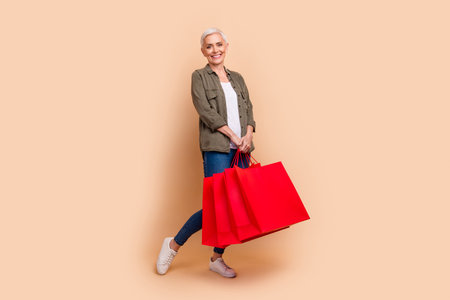 Full body photo of attractive retired woman hold shopping bags dressed stylish khaki clothes isolated on beige color backgroundの写真素材