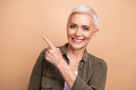 Portrait of mature age white short haircut woman in khaki shirt directing finger mockup lotion advert isolated on beige color backgroundの写真素材