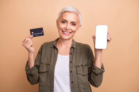 Photo of digital banking features user mature age smiling woman enjoy eshopping hold card and phone...