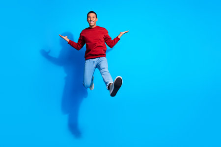 Full size portrait of active cheerful young man jump arms hold empty space isolated on blue color...