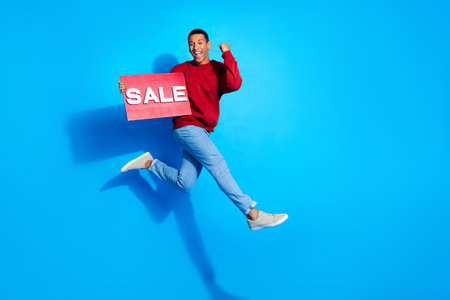 Full size portrait of delighted sporty handsome guy jump run raise fist hold sale placard empty space isolated on blue color backgroundの写真素材