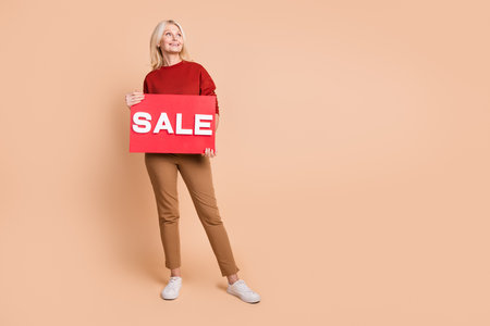 Full length photo of funky dreamy lady dressed red pullover holding sale card looking empty space isolated beige color backgroundの写真素材