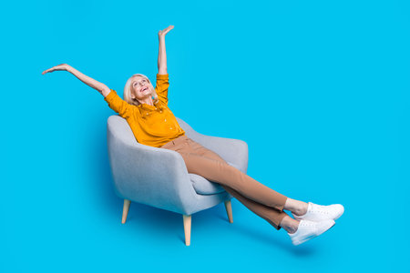 Full length photo of lovely senior lady armchair look up empty space dressed stylish yellow satin garment isolated on blue color backgroundの写真素材
