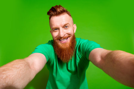 Photo of optimistic funky man with ginger beard dressed green t-shirt making selfie on smartphone isolated on green color backgroundの写真素材