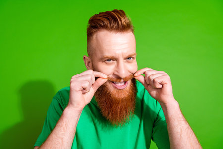 Portrait of funny optimistic guy with red beard dressed green t-shirt touch mustache at barbershop isolated on green color backgroundの写真素材