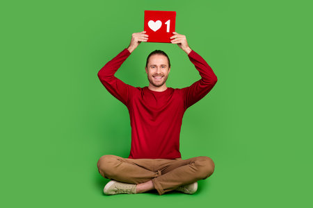 Full size photo good mood handsome guy wear red shirt sit holding red social media like over head isolated on green color backgroundの写真素材