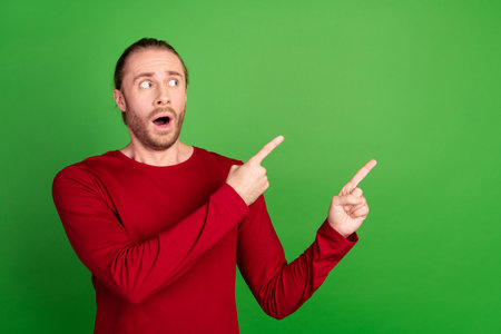 Photo of astonished man with stubble dressed red pullover staring indicating at discount empty space isolated on green color backgroundの写真素材
