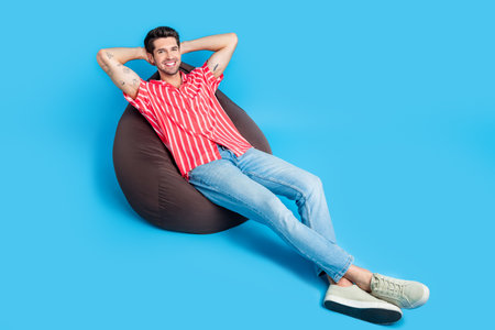 Full size photo of good mood man dressed striped shirt laying on bean bag holding arms behind head isolated on blue color backgroundの写真素材