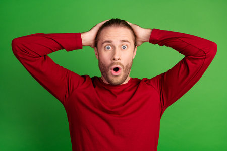 Photo of speechless man with stubble long hairdo dressed red pullover astonished staring arms on head isolated on green color backgroundの写真素材