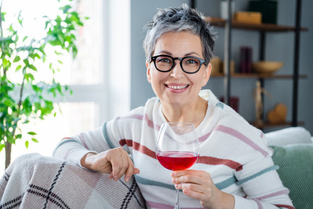 Portrait of cheerful pretty aged person sitting sofa hold red wine glass spend free time modern apartment insideの写真素材