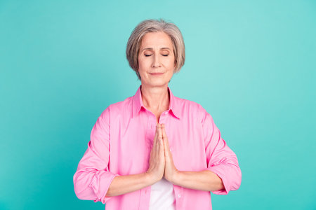 Portrait of relaxing woman with white gray hairdo dressed stylish shirt meditating palms together isolated on teal color backgroundの写真素材