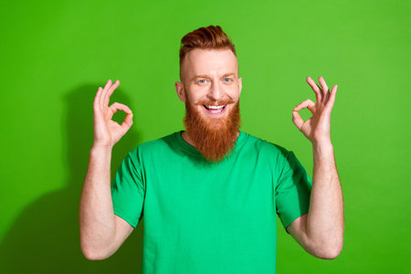 Photo of positive handsome man with ginger long beard dressed stylish t-shirt showing okey good work isolated on green color backgroundの写真素材