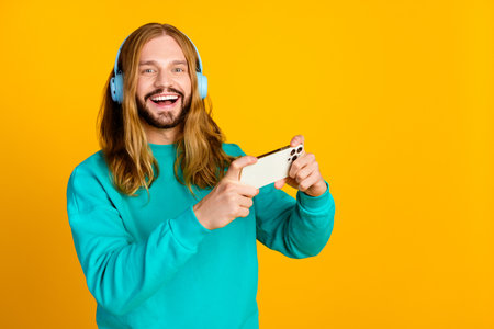 Photo of positive handsome guy listen music headphones play games smart phone empty space isolated...