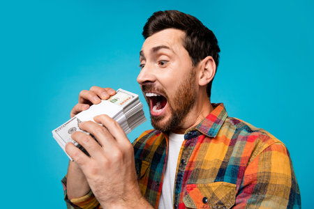 Photo of crazy eccentric man with beard dressed stylish plaid shirt look at dollar banknotes eat money isolated on blue color backgroundの写真素材