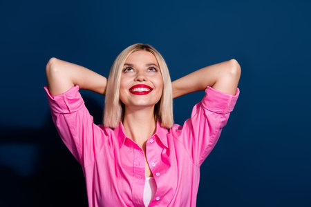 Photo of lovely girl dressed pink shirt holding palms behind head look up at offer empty space isolated on dark blue color backgroundの写真素材