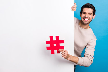 Photo of good mood positive guy dressed beige sweatshirt holding hash tag placard empty space isolated blue color backgroundの写真素材