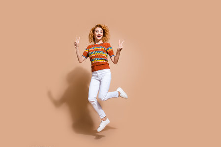 Photo portrait of lovely young lady jumping v-sign dressed stylish striped garment isolated on beige...