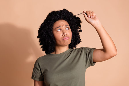 Photo of disappointed woman with afro hairstyle wear khaki t-shirt look at curl dislike new hairdo isolated on beige color backgroundの写真素材