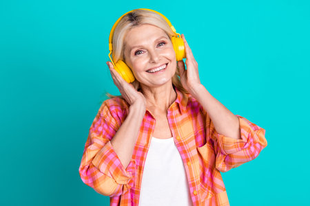 Photo of good mood senior person with blond hairstyle dressed checkered shirt arms touching headphones isolated on teal color backgroundの写真素材