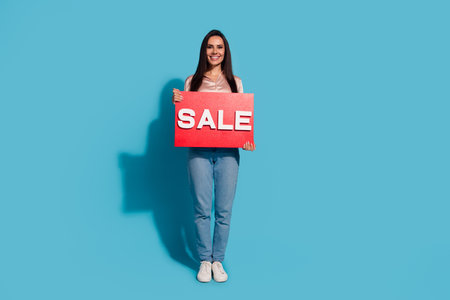 Full length photo of positive cheerful adorable girl wear silk blouse jeans hold red sale placard solated on blue color backgroundの写真素材