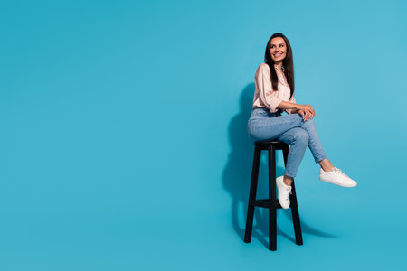 Full length photo of good mood thoughtful lady dressed beige shirt sitting chair looking empty space isolated blue color backgroundの写真素材