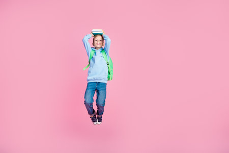 Full size portrait of cheerful schoolchild jump hold book above head empty space ad isolated on pink...