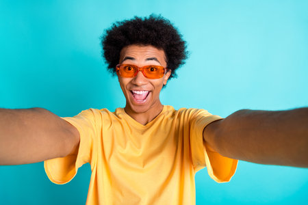 Photo of overjoyed funky guy with afro hair dressed yellow t-shirt in sunglass making selfie isolated on turquoise color backgroundの写真素材
