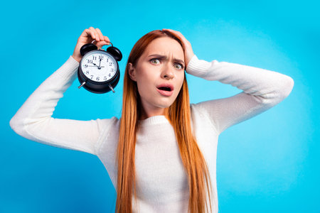 Photo portrait of attractive young woman hold alarm clock confused dressed stylish white clothes isolated on blue color backgroundの写真素材