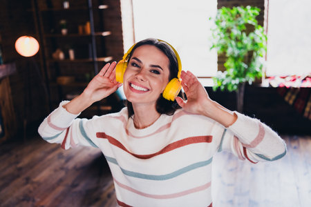 Photo of lovely young woman dancing listen music headphones wear striped outfit interior home living room in brown warm colorの写真素材