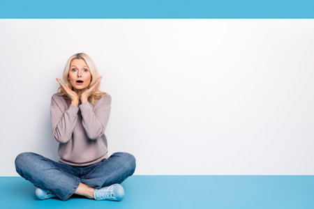 Full body photo of senior lady sit floor crossed legs empty space afraid dressed stylish gray garment isolated on blue color backgroundの写真素材
