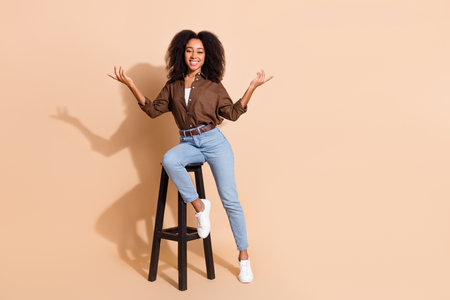 Full size photo of positive woman dressed brown shirt sit on chair arms presenting products empty space isolated on beige color backgroundの写真素材