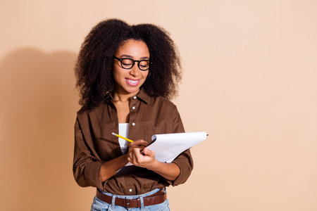 Portrait of clever woman with wavy hair dressed brown shirt in eyewear write in clipboard at...