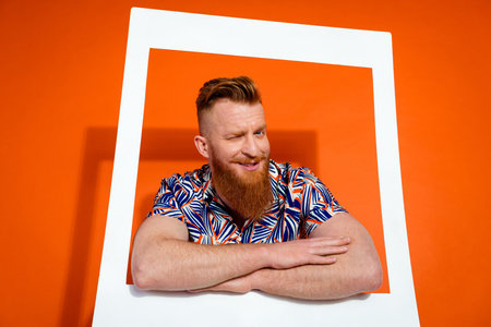 Photo of optimistic guy with red beard wear print shirt hold hands crossed in frame blinking eye isolated on orange color backgroundの写真素材