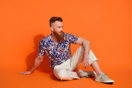 Full length photo of brutal guy wear print shirt trousers sit on floor look at offer empty space isolated on orange color backgroundの写真素材