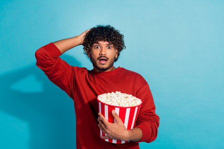 Photo of shocked speechless impressed reaction young guy in red pullover touch head staring with popcorn isolated on cyan color backgroundの写真素材
