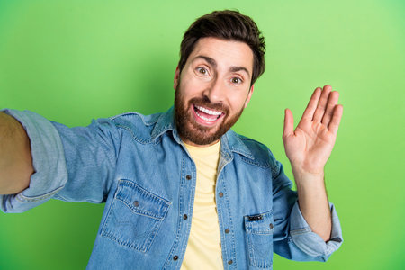 Photo of good mood handsome guy wear denim jacket recording video wacing arm palm hi empty space isolated green color backgroundの写真素材