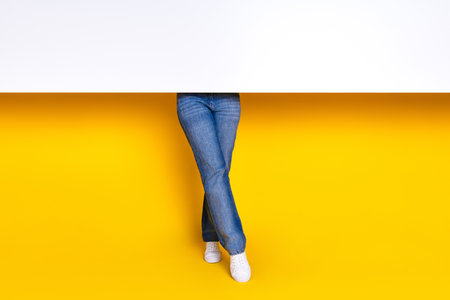 Cropped photo of stylish woman crossed legs dressed denim pants stand behind white placard empty space isolated on yellow color backgroundの写真素材