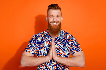 Photo of cheerful positive guy dressed print shirt arms together praying isolated orange color backgroundの写真素材