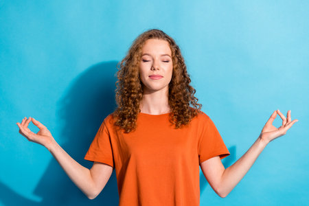 Portrait of youngster cheerful girl with beautiful curly red hair wearing orange t shirt balance in harmony isolated on blue color backgroundの写真素材