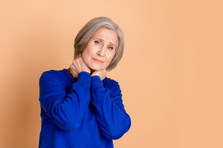 Photo portrait of lovely pensioner lady throat pain suffer dressed stylish blue garment isolated on beige color backgroundの写真素材