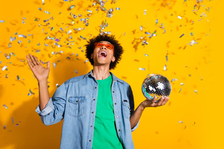 Photo of nice young man disco ball confetti wear denim shirt isolated on yellow color backgroundの写真素材