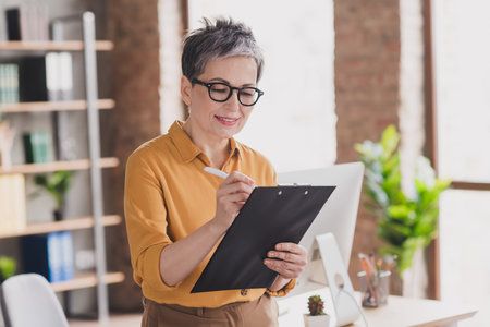 Photo of lovely mature lady signing documents dressed yellow formalwear modern workplace success business owner home officeの写真素材