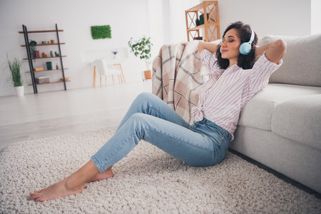 Full length photo of shiny cute girl dressed white striped shirt headphones arms behind head enjoying songs indoors house apartment roomの写真素材