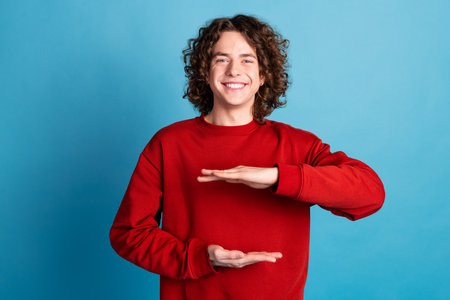 Photo of attractive funny guy wear red sweater showing arms height emtpy space isolated blue color backgroundの写真素材