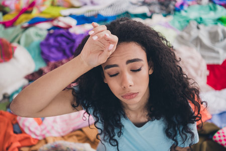 High angle view photo of young girl hand touch forehead pile stack clutter clothes apartment indoorsの写真素材
