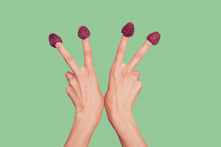 Close-up cropped delicate well-groomed two hands four fingers sweet berries hats on nails showing v-sign isolated over pink pastel backgroundの写真素材