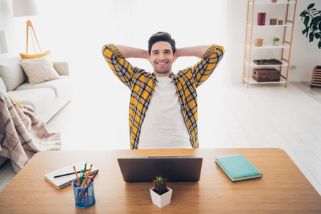 Photo of cheerful positive guy dressed plaid shirt arms behind head working modern gadget indoors room home houseの写真素材