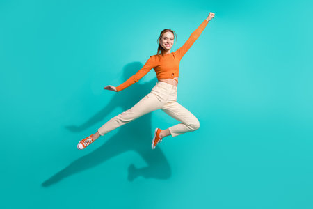Full size photo of adorable cool girl dressed orange shirt pants flying empty space holding fist up isolated on teal color backgroundの写真素材