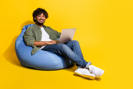 Photo of young arab guy remote studying in khaki shirt sitting pouf junior it specialist at lounge isolated on yellow color backgroundの写真素材
