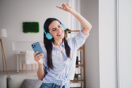 Portrait of pretty lady smart phone headphones listen music wear shirt bright interior apartment indoorsの写真素材